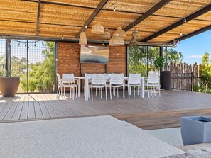 An outdoor patio with a wooden deck, white chairs, a table, and a TV screen. - 10 Bedroom / Pool / Hot tub / Pickleball / Firepit (Dripping Springs)