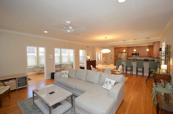 A spacious living room with a large grey sectional sofa, a coffee table, and a dining area with a round table and chairs. A kitchen with wooden cabinets and a bar area is visible in the background.