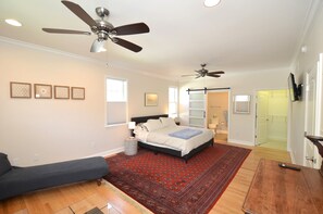 A bedroom with a bed, two ceiling fans, a red patterned rug, a wooden floor, and a small table with a remote control.