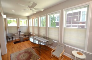 A spacious room with a round glass table, two chairs, a small white table, and a ceiling fan.