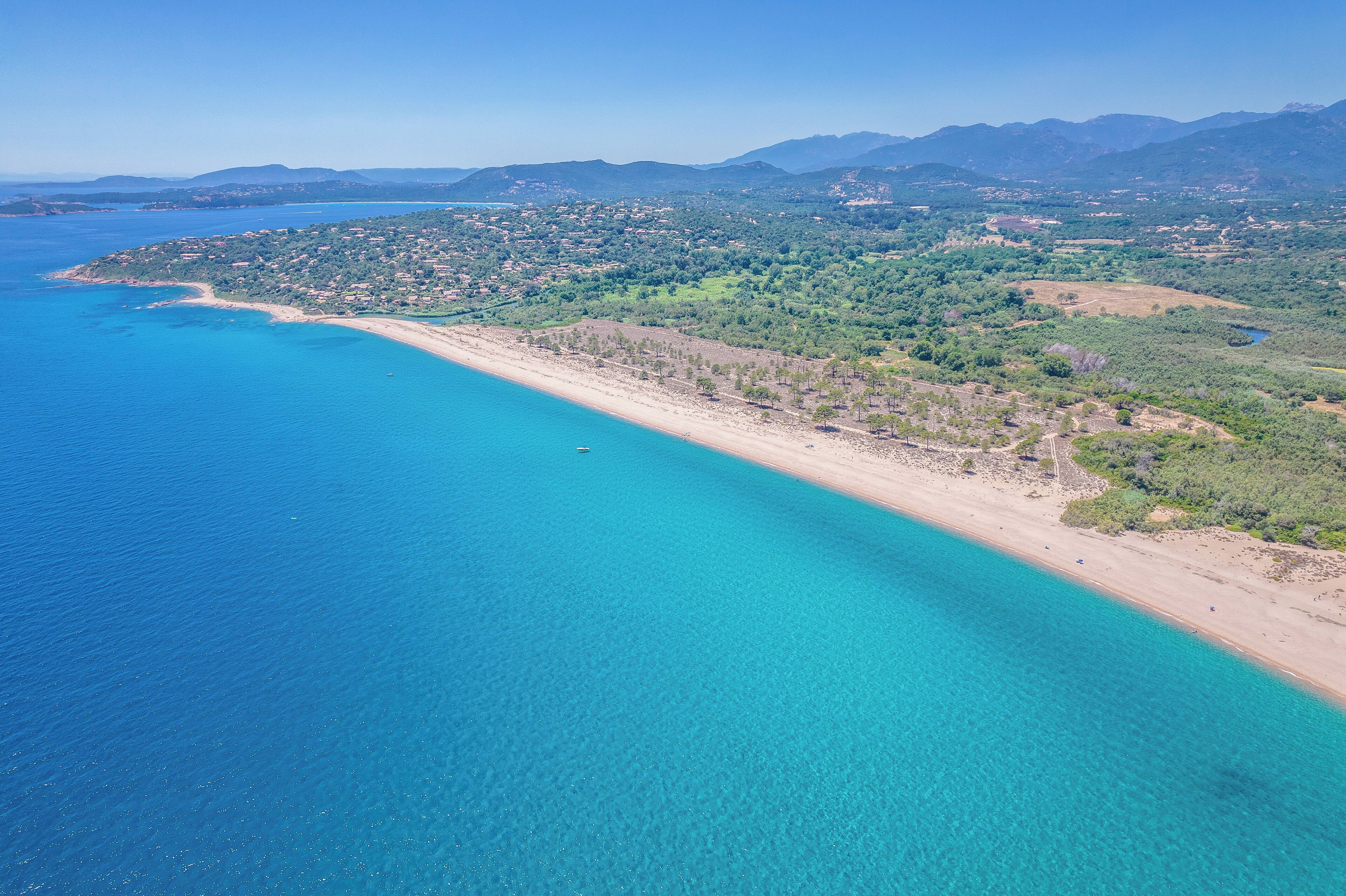 Luftaufnahme einer Küstenlandschaft mit Sandstrand, klarem, blauem Wasser und einer Bergkulisse im Hintergrund.