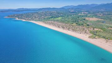 Luftaufnahme einer Küstenlandschaft mit Sandstrand, klarem, blauem Wasser und einer Bergkulisse im Hintergrund.