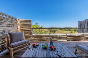 Une terrasse en bois avec une table dressée pour deux personnes, agrémentée d’un saladier de raisins verts, de tranches de pastèque et d’un vase.