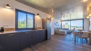Une cuisine moderne avec des meubles sombres, un grand îlot central et un réfrigérateur encastré. On y trouve un coin repas avec une table et des chaises, ainsi qu'un canapé. De grandes fenêtres laissent entrer la lumière naturelle et offrent une vue sur l'extérieur.