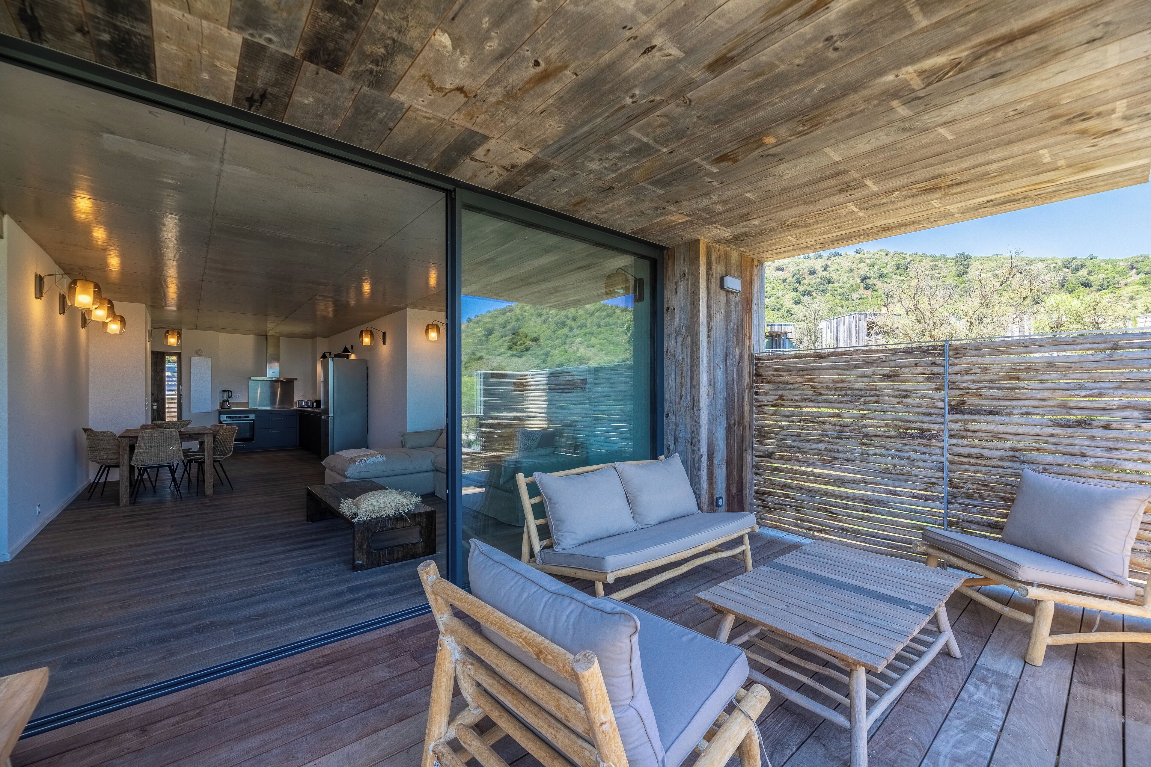 Un espace de vie extérieur moderne, doté de mobilier en bois, d’un mur en verre et offrant une vue sur une colline verdoyante.