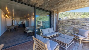Un espace de vie extérieur moderne, doté de mobilier en bois, d’un mur en verre et offrant une vue sur une colline verdoyante.