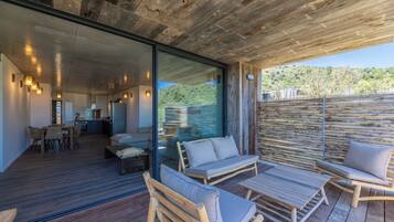 Un espace de vie extérieur moderne, doté de mobilier en bois, d’un mur en verre et offrant une vue sur une colline verdoyante.