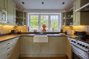 A kitchen with light-colored cabinets, a double sink, a stove, and a window with a view of greenery outside.