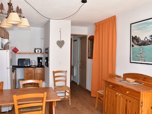 Cucina compatta con forno a microonde, tostapane e macchina del caffè. Angolo ristoro con tavolo e sedie in legno, e un piccolo mobile con cassetti. Sul muro, una cornice con un'immagine del Parco Nazionale della Vanoise.