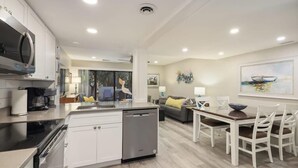 Una cocina moderna con armarios blancos, lavavajillas y horno empotrado. Hay una zona de comedor con mesa de madera y sillas blancas.