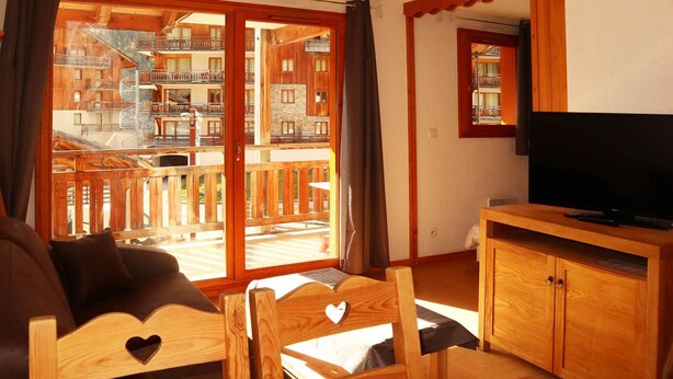 A cozy living room with a wooden sofa, a TV on a wooden stand, and a large window with a view of a building with balconies. - Ski-in/ski-out apartment with indoor pool, close to amenities (Les Orres)