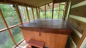 Un bain à remous en bois, doté d’un banc, de grandes fenêtres et offrant une vue sur la verdure.