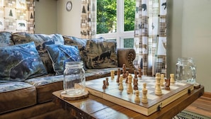 Un salon chaleureux avec un jeu d’échecs posé sur une table en bois, un canapé agrémenté de coussins à motifs et une fenêtre avec des rideaux.