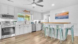 A modern kitchen with white cabinets, stainless steel appliances, and a bar area with green stools. - Poolside Paradise: Modern Coastal Oasis with Private Backyard Pool (Oak Island)