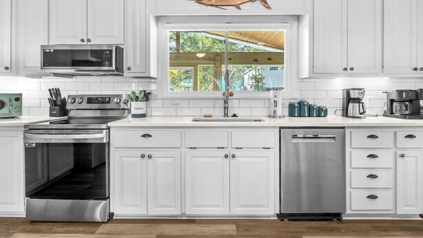 A kitchen with white cabinets, stainless steel appliances, a microwave, oven, and dishwasher. - Poolside Paradise: Modern Coastal Oasis with Private Backyard Pool (Oak Island)