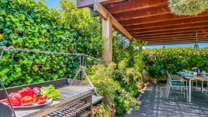 Une terrasse extérieure avec un barbecue, une table dressée pour les repas et une végétation luxuriante qui entoure l’espace.