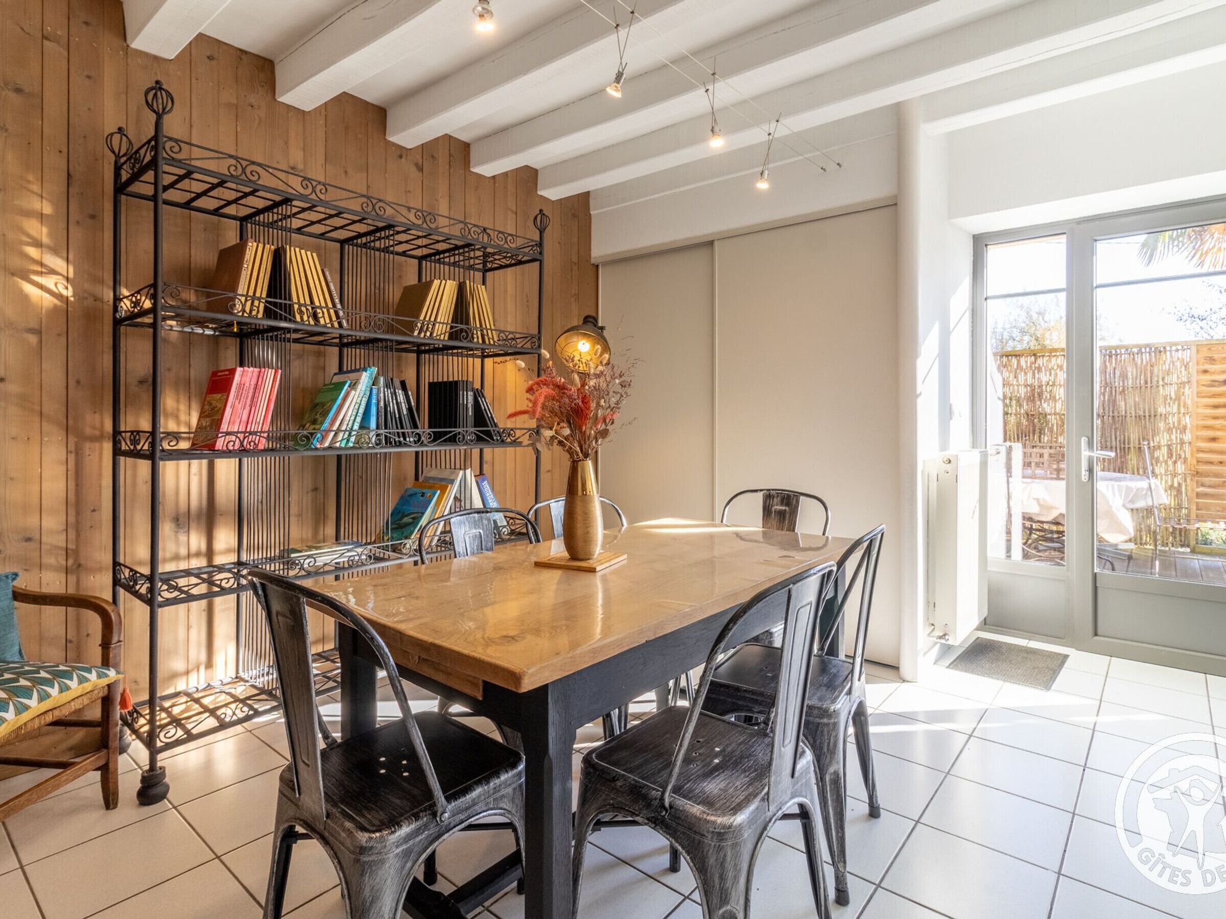 Zona pranzo con tavolo e sedie in legno, una libreria metallica con libri e un vaso con fiori secchi.