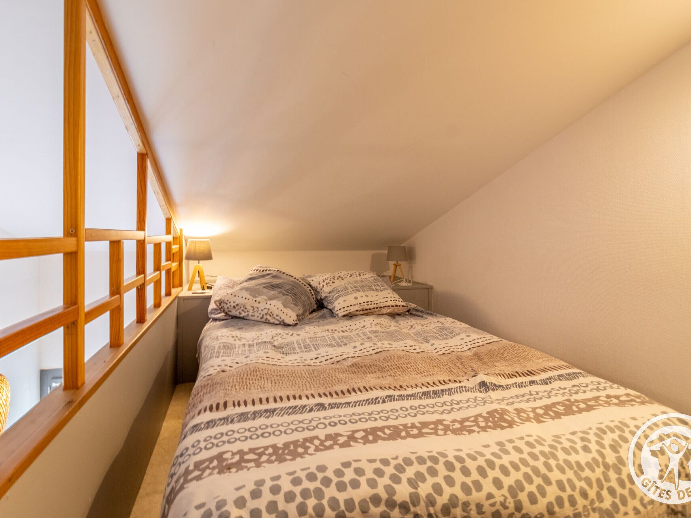 A bedroom with a sloped ceiling, a bed with patterned bedding, two bedside lamps, and a window with wooden frames.