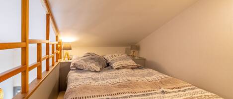 A bedroom with a sloped ceiling, a bed with patterned bedding, two bedside lamps, and a window with wooden frames.