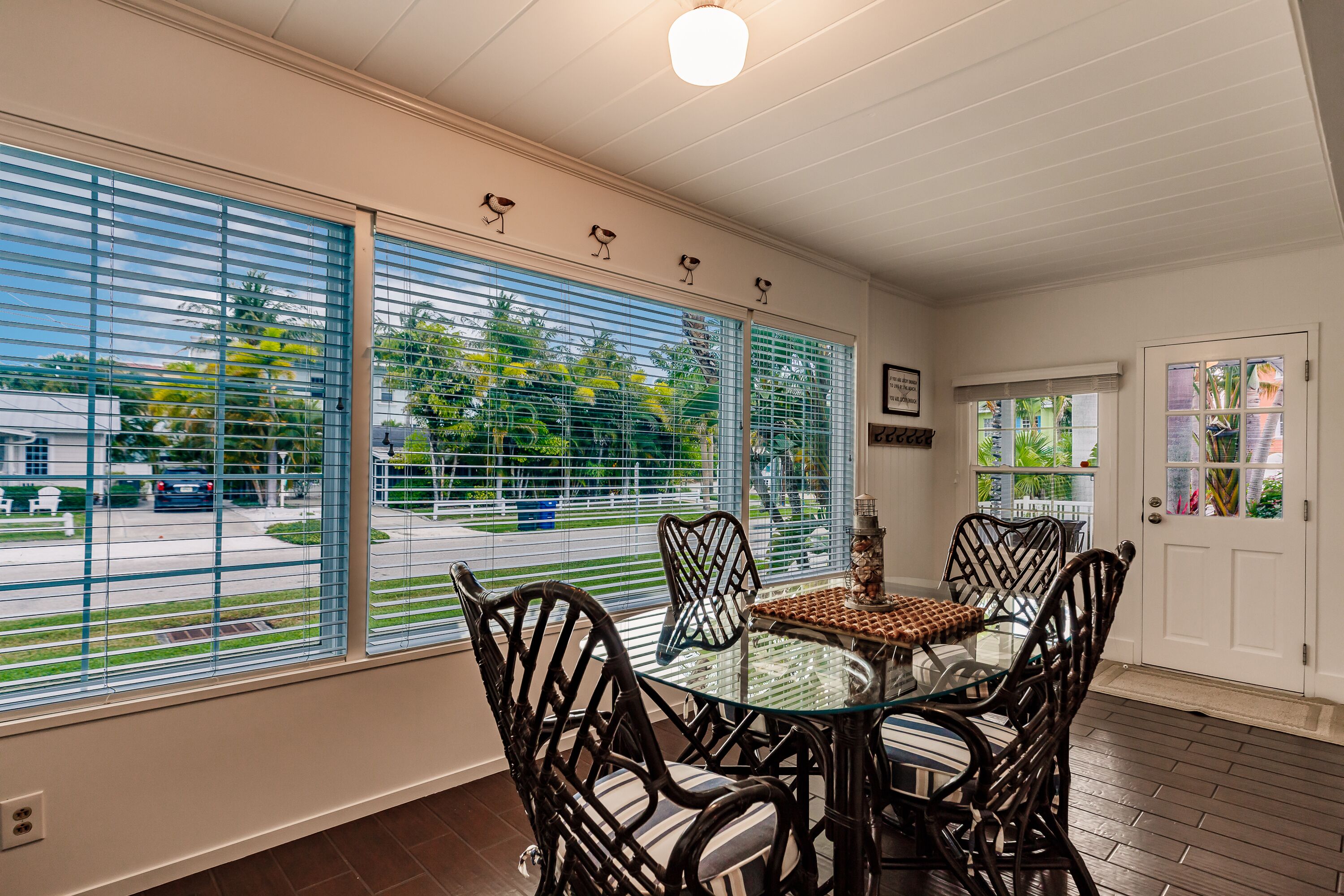 A dining area with a glass table and wicker chairs, large windows with blinds, and a white door with a glass panel.