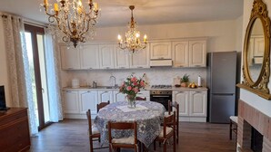 A dining area with a round table, chairs, a chandelier, and a kitchen in the background. - Villa (Reggello)