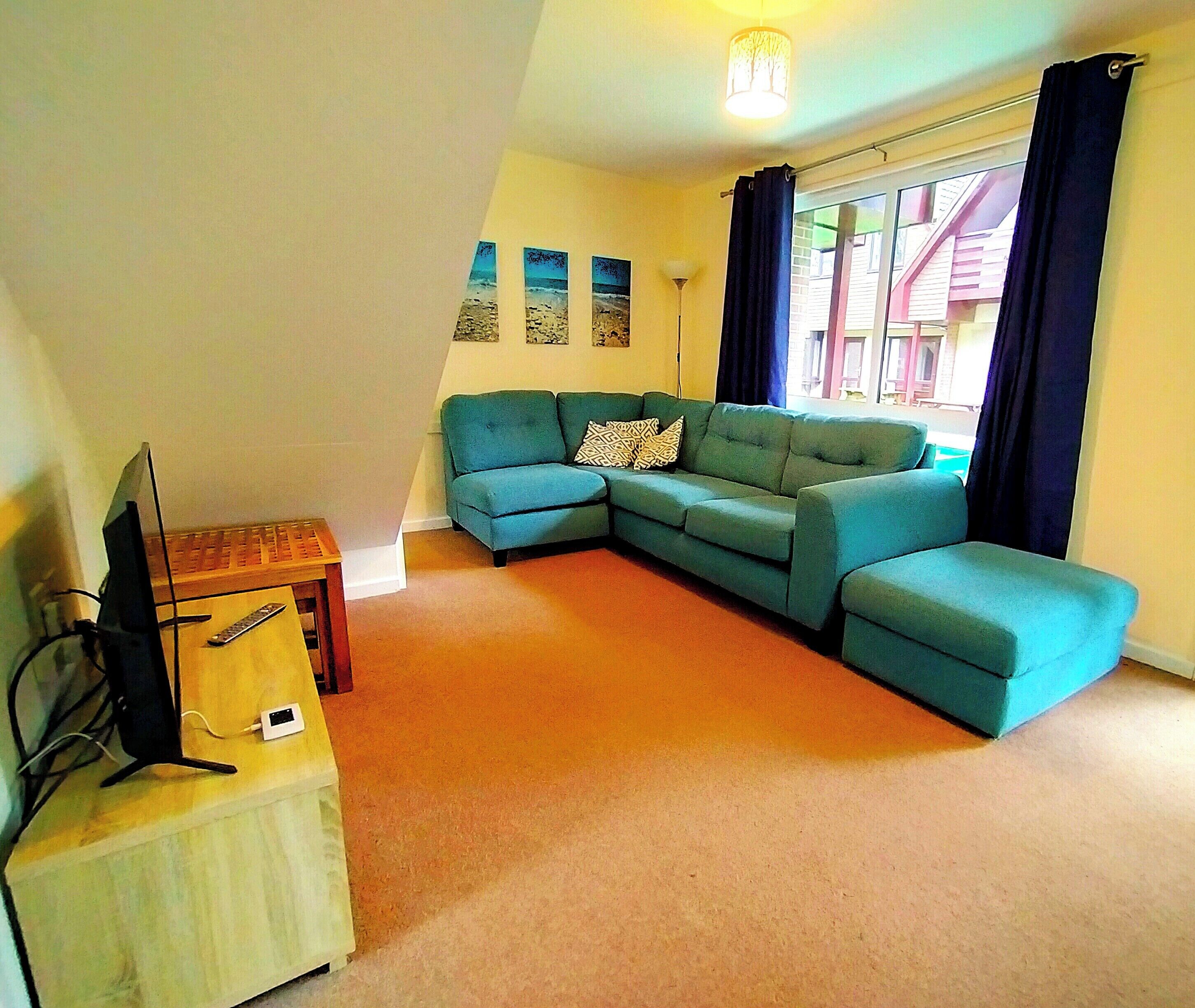 A living room with a teal sofa, a television on a wooden stand, and a window with curtains.