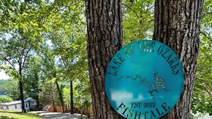 A sign on a tree reading "Lake of the Ozarks Est. 2022 Fish Tale." - Updated Ozarks Lakefront Home-Lilly pad, kayaks, Private dock & boat ramp. (Sunrise Beach)