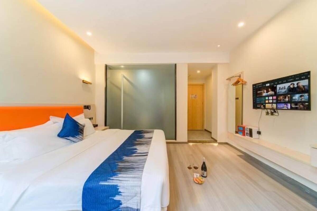 Habitación de hotel moderna con una cama grande, una puerta de vidrio que da a un pasillo y un televisor con pantalla montado en la pared mostrando varios canales.