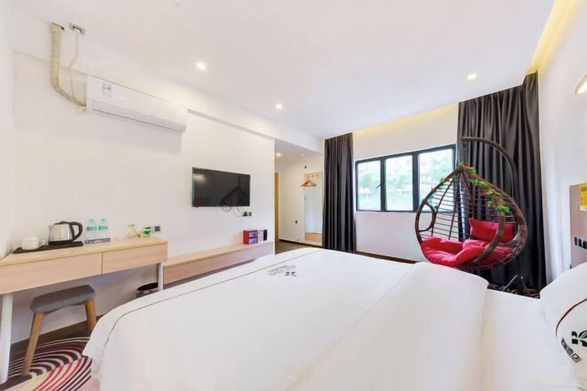 Habitación de hotel con cama, escritorio con silla, televisor montado en la pared y sillón colgante con cojines rojos.