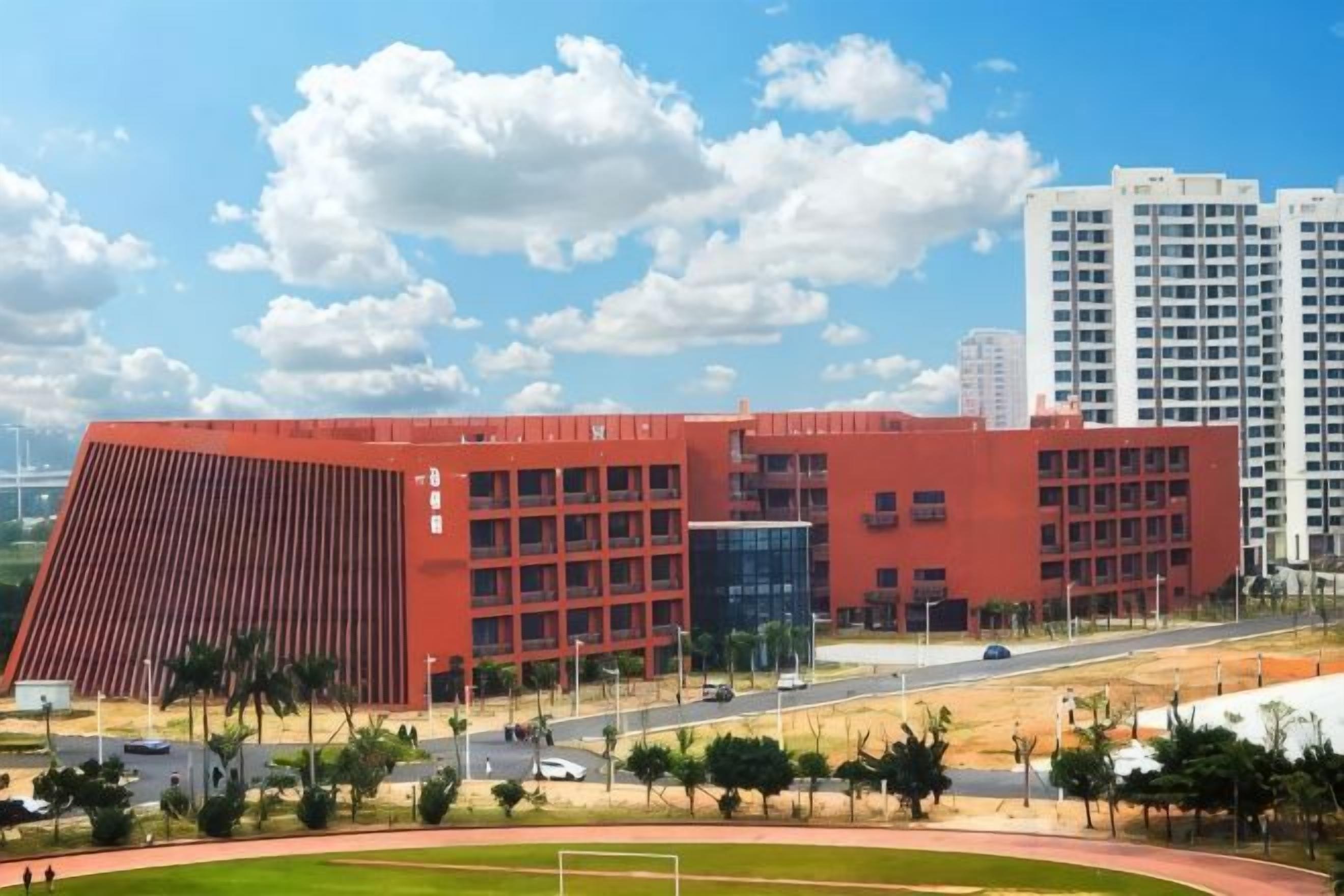 A large red building with multiple floors, surrounded by a landscaped area with trees and a road.