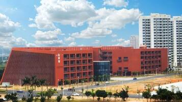 A large red building with multiple floors, surrounded by a landscaped area with trees and a road.