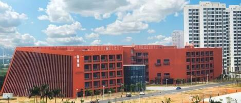 Un edificio rojo grande con varios pisos, rodeado de una zona ajardinada con árboles y una calle.