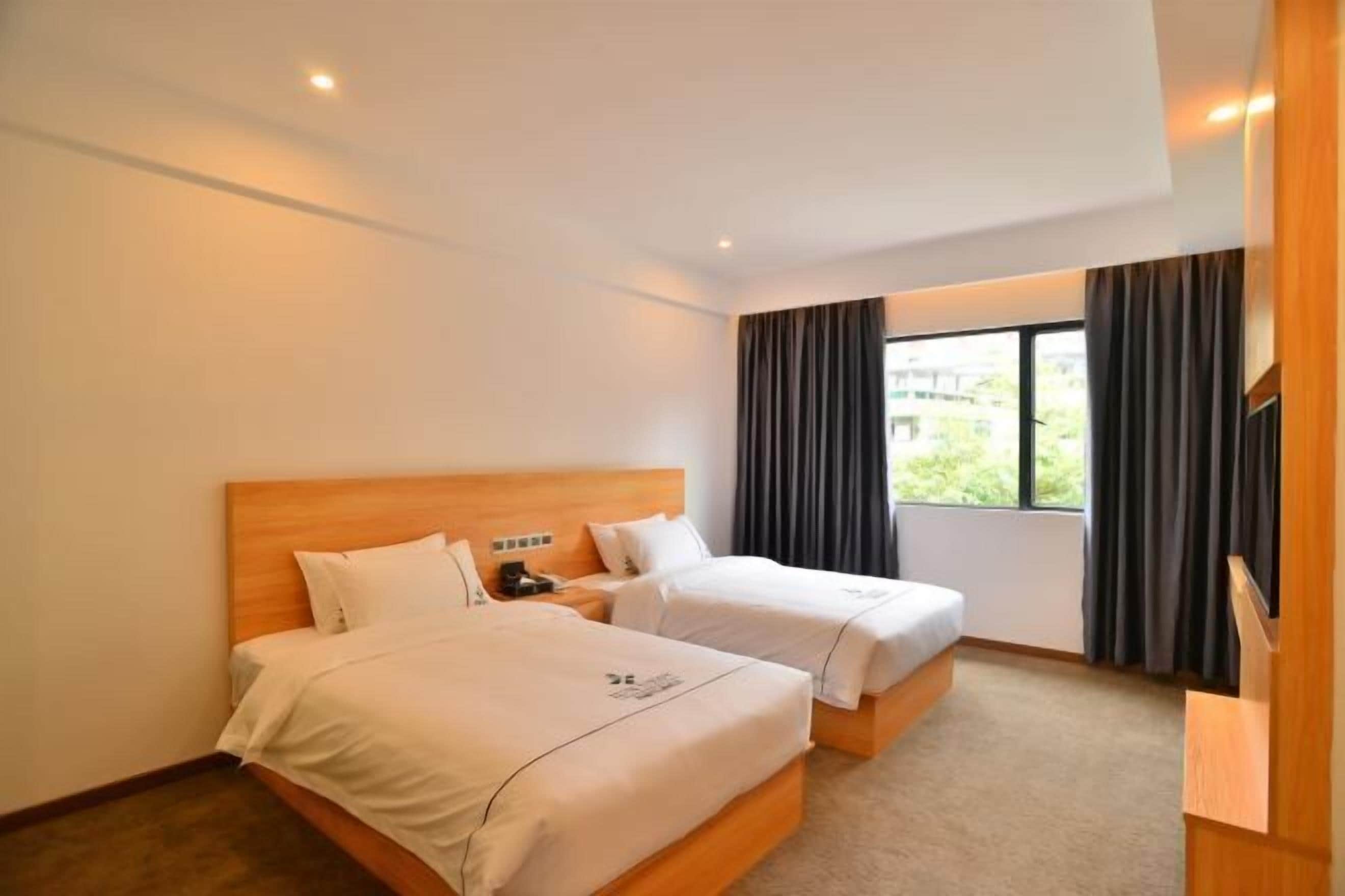 A hotel room with two beds, a wooden headboard, a nightstand, a window with curtains, and a view of greenery outside.