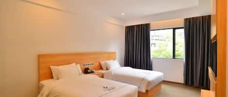 A hotel room with two beds, a wooden headboard, a nightstand, a window with curtains, and a view of greenery outside.