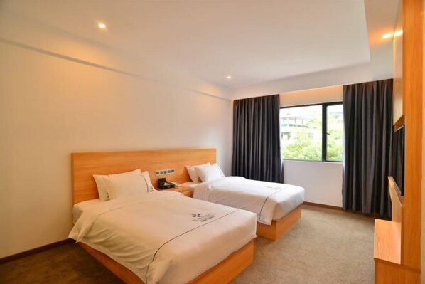A hotel room with two beds, a wooden headboard, a nightstand, a window with curtains, and a view of greenery outside. - Junchao Light Luxury Hotel (Zhuhai)