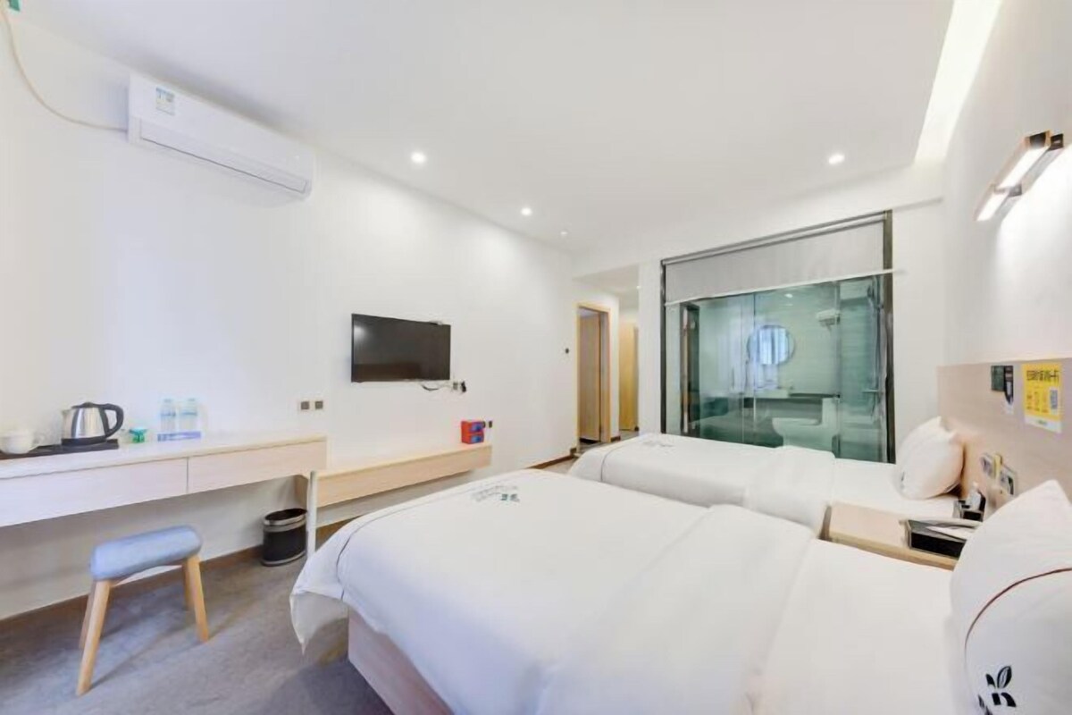 A modern hotel room with a desk, two beds, a TV, and a glass shower area.