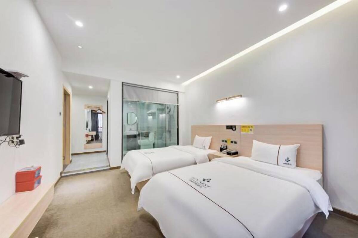 Habitación de hotel con dos camas, televisión, escritorio y puerta de vidrio que da al baño.