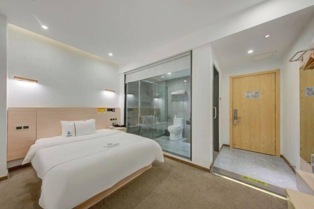 Habitación de hotel con cama, puerta de madera, un separador de vidrio y un baño pequeño visible a través del vidrio.