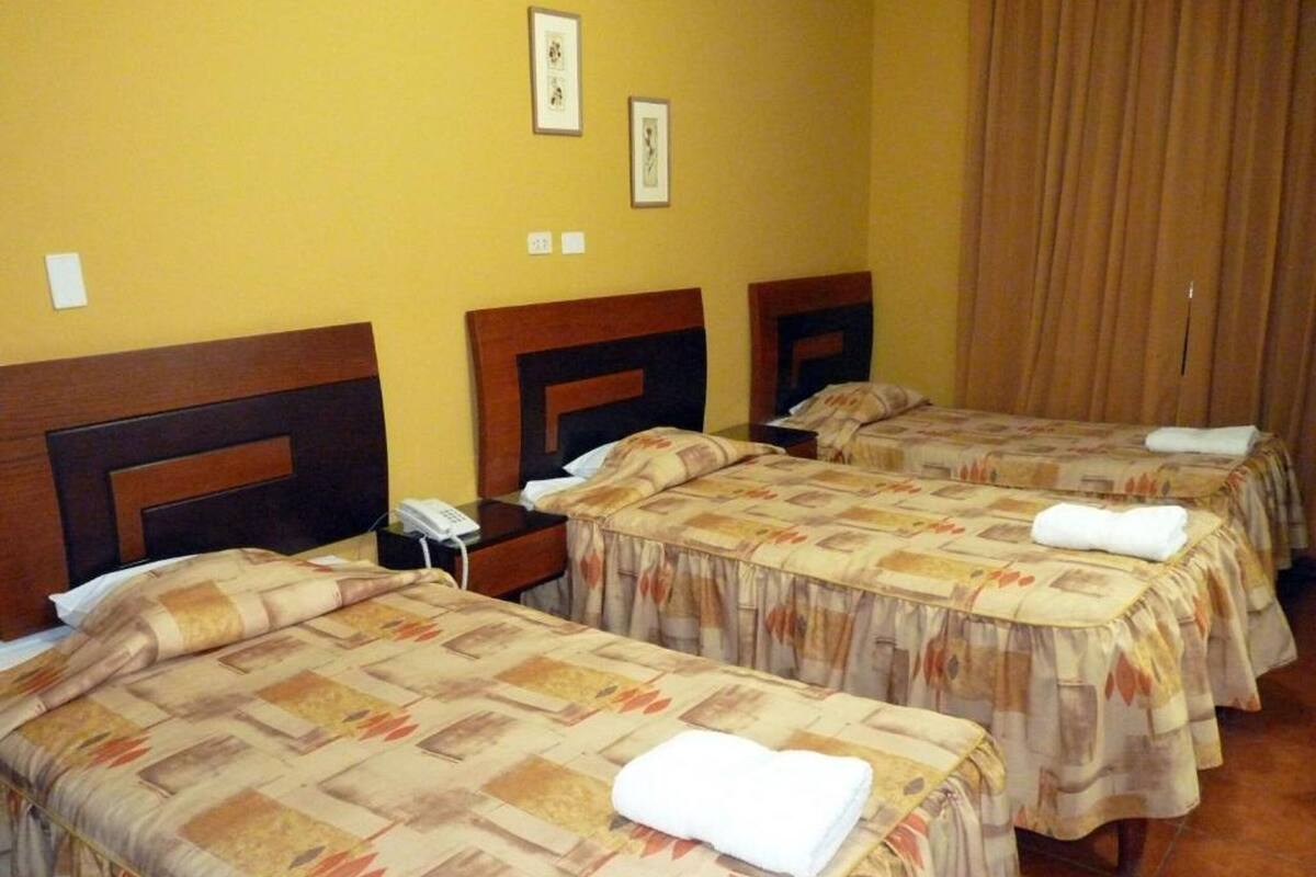 A hotel room with three beds, each with a patterned bedspread and a white towel folded on top. There are bedside tables with a telephone and a lamp. The walls are yellow, and there are framed pictures on the wall.