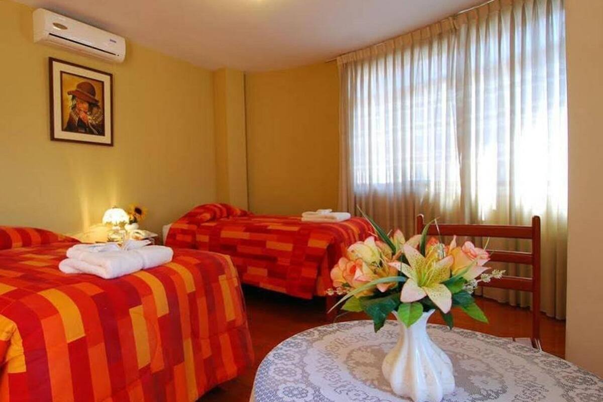 A hotel room with two beds, a bedside table with a lamp, a vase with flowers, and a window with curtains.