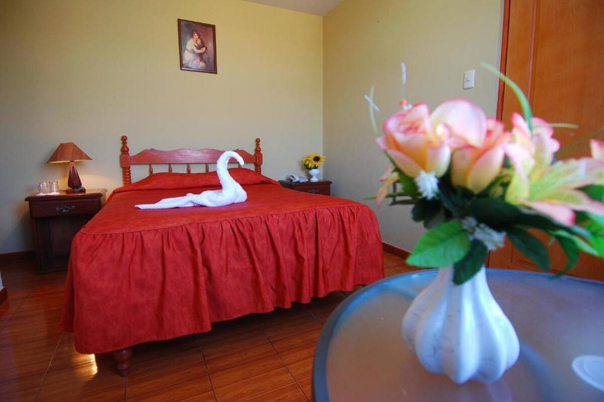 A hotel room with a red bedspread, a wooden headboard, a nightstand with a lamp, a vase with flowers, and a framed picture on the wall.