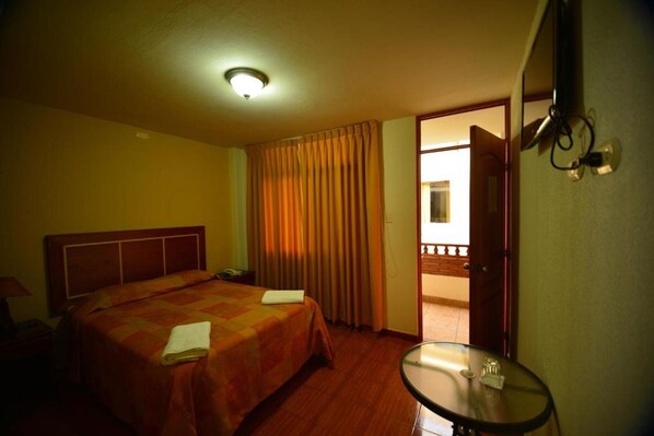 A hotel room with a bed, a small table, a door leading to a balcony, and a ceiling light.