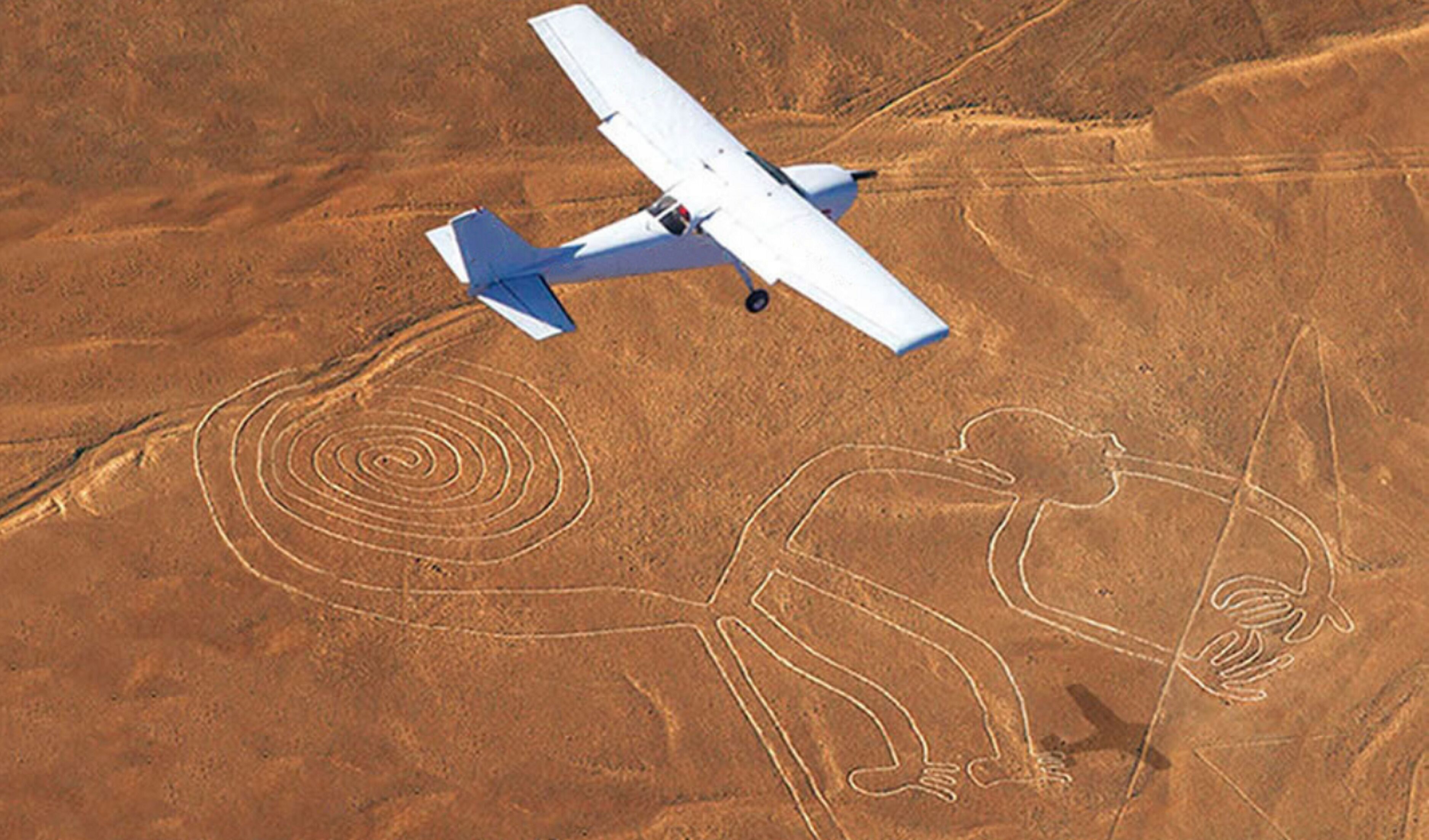 Un avión vuela sobre un paisaje desértico con figuras en espiral y figuras que parecen humanas grabadas en el suelo.