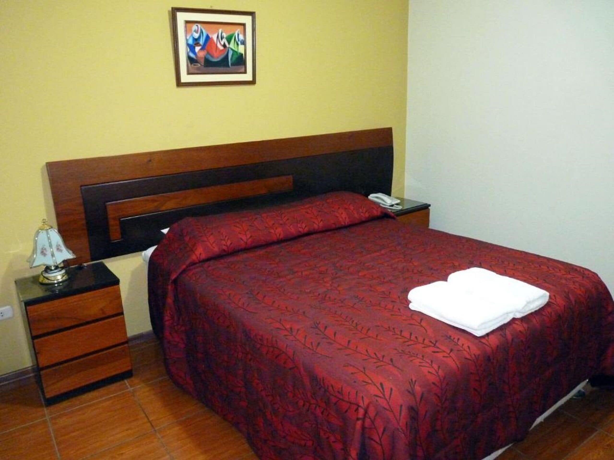 Una cama bien hecha con una colcha roja estampada y una toalla blanca doblada encima. Hay una mesita de noche con una lámpara y un teléfono.