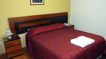Una cama bien hecha con una colcha roja estampada y una toalla blanca doblada encima. Hay una mesita de noche con una lámpara y un teléfono.