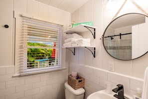 A bathroom with white tiles, a toilet, a sink, a mirror, and a window with blinds. - Cozy Cottage with Hot Tub (Siesta Key)