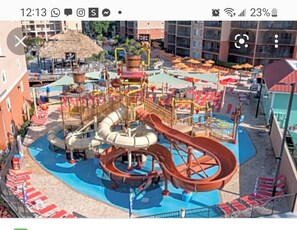 Enjoy the resort's water park with slides, play structures, and a pool area surrounded by lounge chairs and a thatched-roof pavilion.