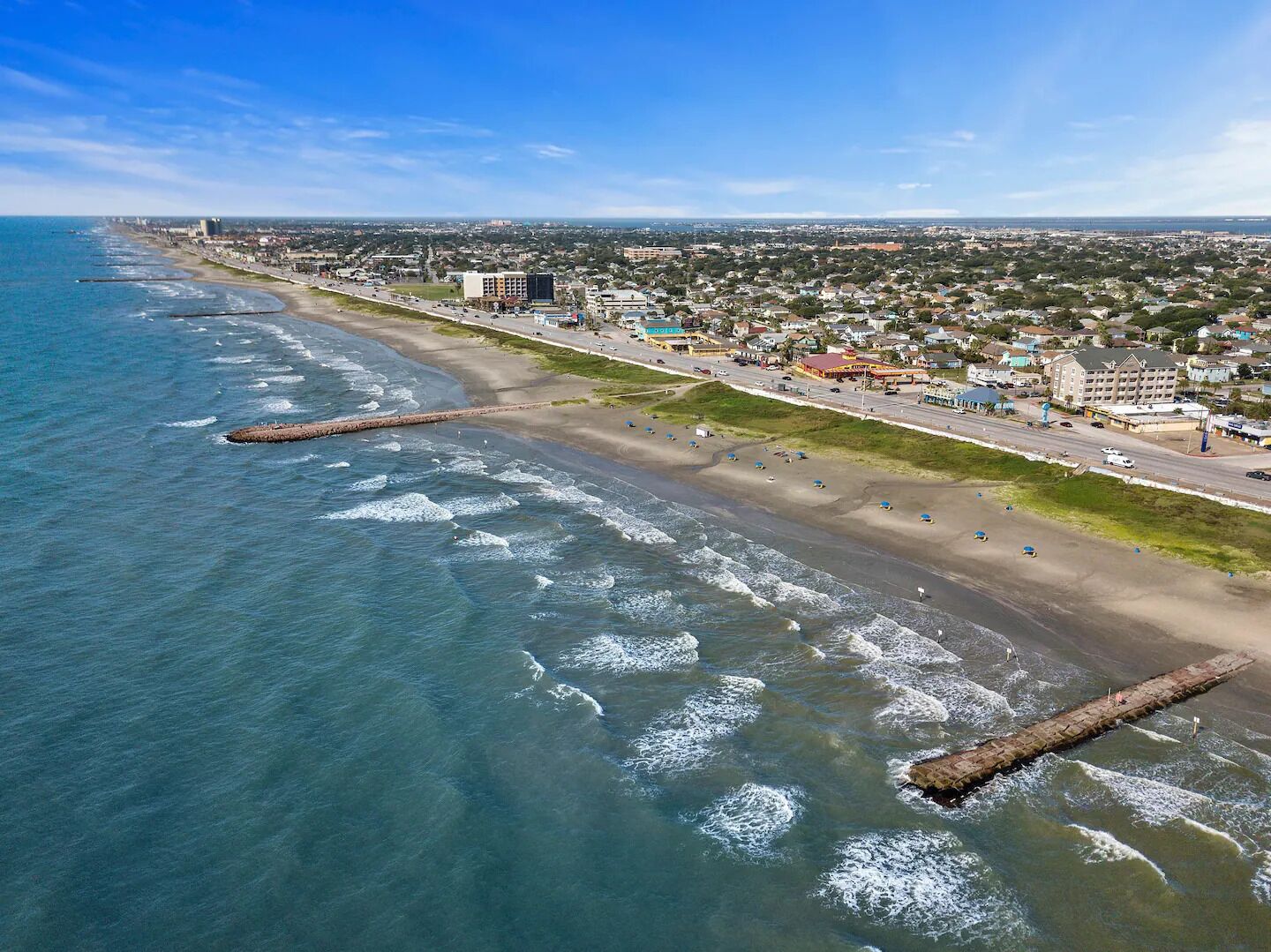An aerial view of a coastal city with a beach, buildings, and a road.