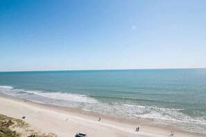 Una playa con cielo despejado, mar tranquilo y algunas personas caminando por la orilla.