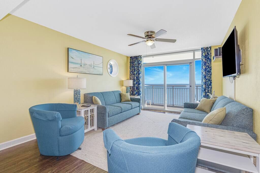 A living room with a ceiling fan, a large window with a view, and a flat-screen TV mounted on the wall.